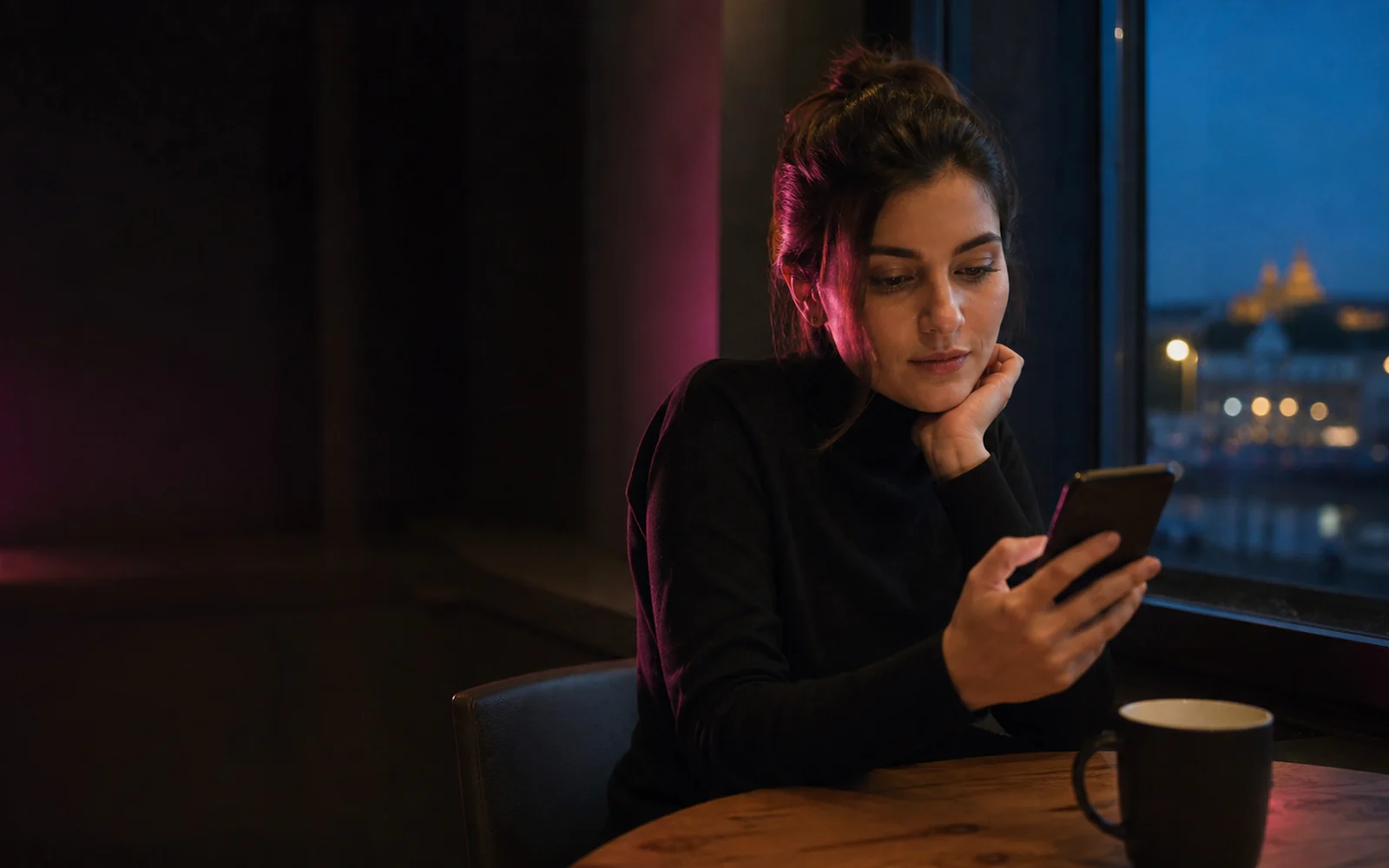 Nő elgondolkodva olvas egy üzenetet a telefonján egy esti kávézóban